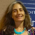 Foto da professora Rebecca Abers sorrindo. A foto foca apenas no rosto e detalhes até o início do tronco. Uma mulher branca, de cabelos grisaçhos na altura dos ombros, soltos com cachos largos no fim dos fios e uma blusa cavada de flores com um colar azul de sementes pintadas.